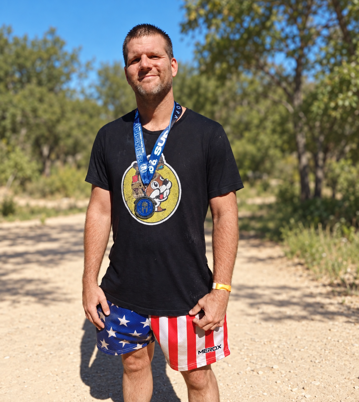 Adult man wearing MEROX USA American flag mesh shorts outdoors; adapted source photo with small-logo branding and a simplified park background.
